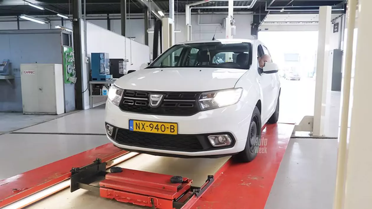 Mașină de culoare albă model Dacia Sandero, ridicată pe o rampă într-un service auto. Se observa numerele de înmatriculare de culoare galbenă și codul de țară al Olandei.