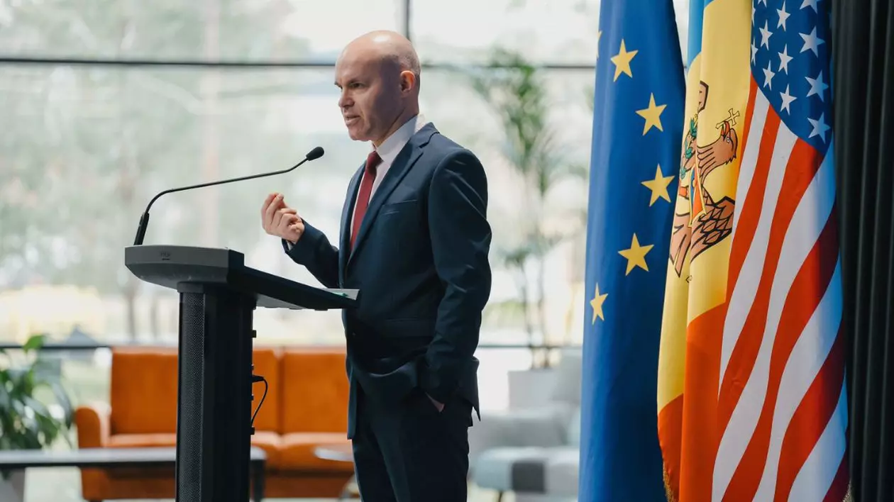 Daniel Funeriu in costum la un pupitru cu microfon avand in spate steagurile UE, Romaniei si SUA