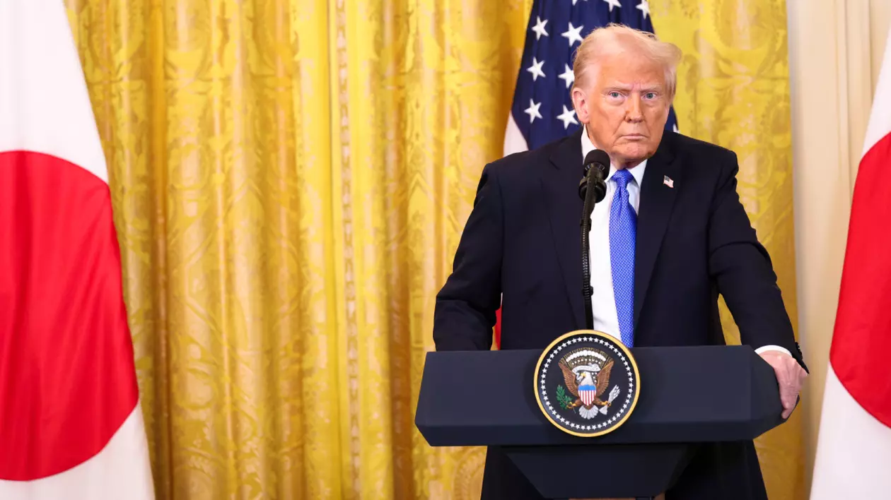 Donald Trump, îmbrăcat într-un costum albastru închis, cu cămașă albă și cravată albastru deschis, stă cu mâinile pe un pupitru care are un microfon. În spatele lui e o cortină galbenă și steagul americii.
