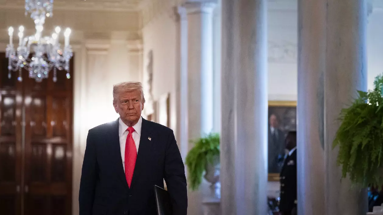 Donald Trump, îmbrăcat într-un costum albastru închis, cămașă albă și cravată roșie, mergând pe un hol. În dreapta lui sunt niste ferestre cu perdele albe, în stânga sus este un candelabru.