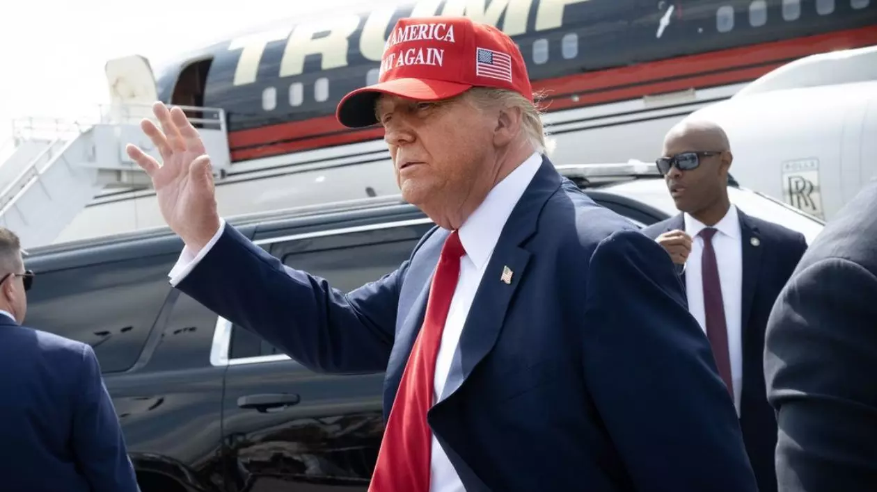 Donald Trump, pe aeropost, lângă un avion, în costum negru cu o cravată roșie si cu o paparie tot rosie, cu mâna dreaptă ridicată