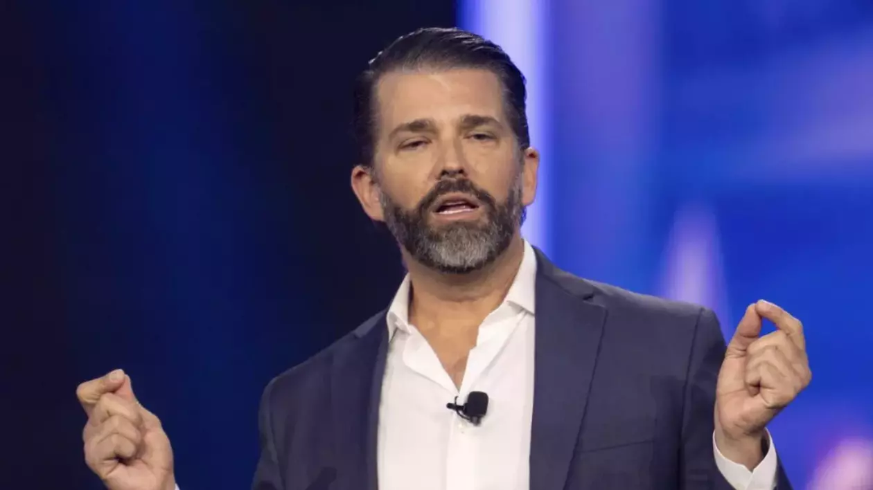În imagine este ilustrat Donald Trump Jr, îmbrăcat într-un costum și cămașă albă, în timp ce vorbește