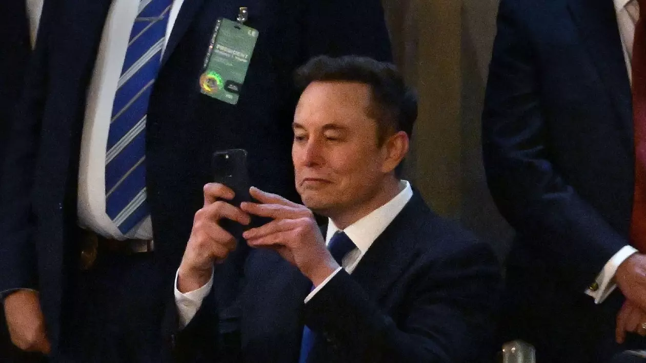 Elon Musk îmbrăcat în costum negru și cămașă albă, cu un telefon în mână