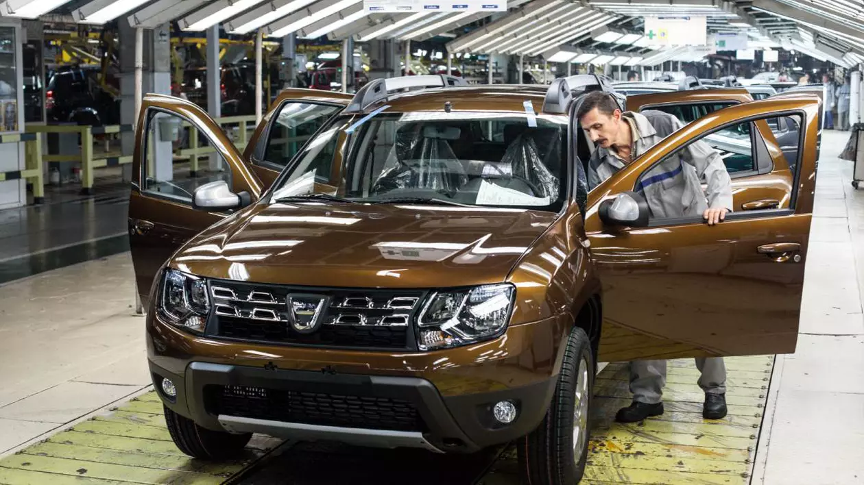 Interiorul unei fabrici auto Dacia din Mioveni, România. În prim-plan se află un model Dacia Duster de culoare maro aflat pe linia de asamblare, în timp ce un muncitor inspectează atent interiorul mașinii.
