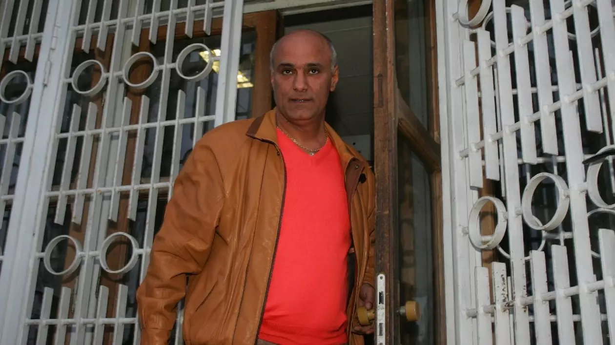 Un bărbat cu geacă maro și tricou portocaliu iese pe o poartă din fier