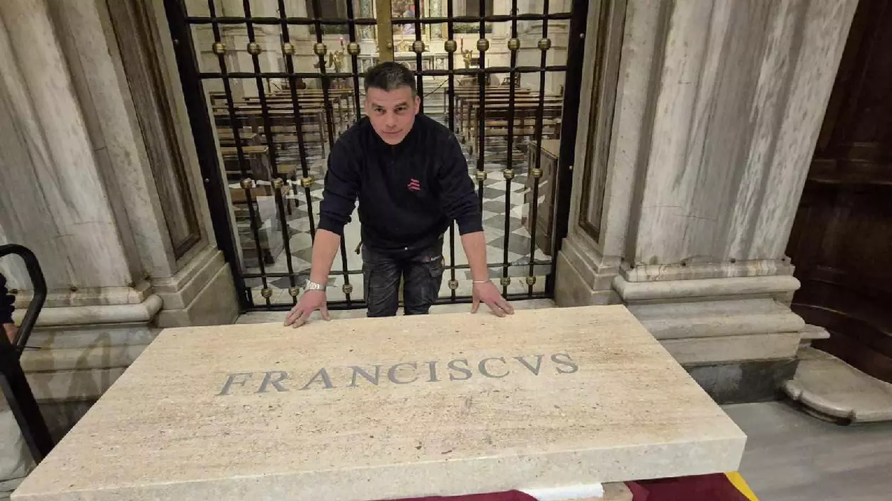 Un bărbat îmbrăcat în negru stă cu mâinile sprijinite pe o placă de marmură pe care este inscripționat „Franciscus”