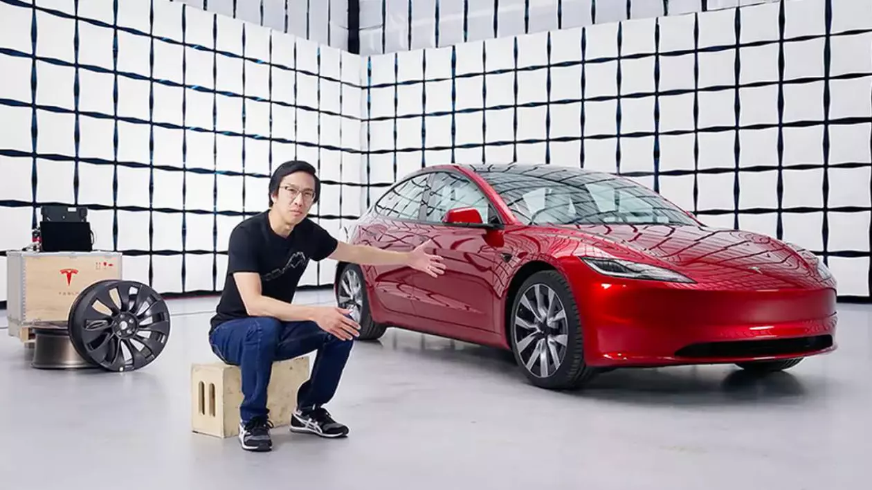 un barbat care sta asezat pe un cub, imbracat in negru, in spatele lui este o masina tesla rosie