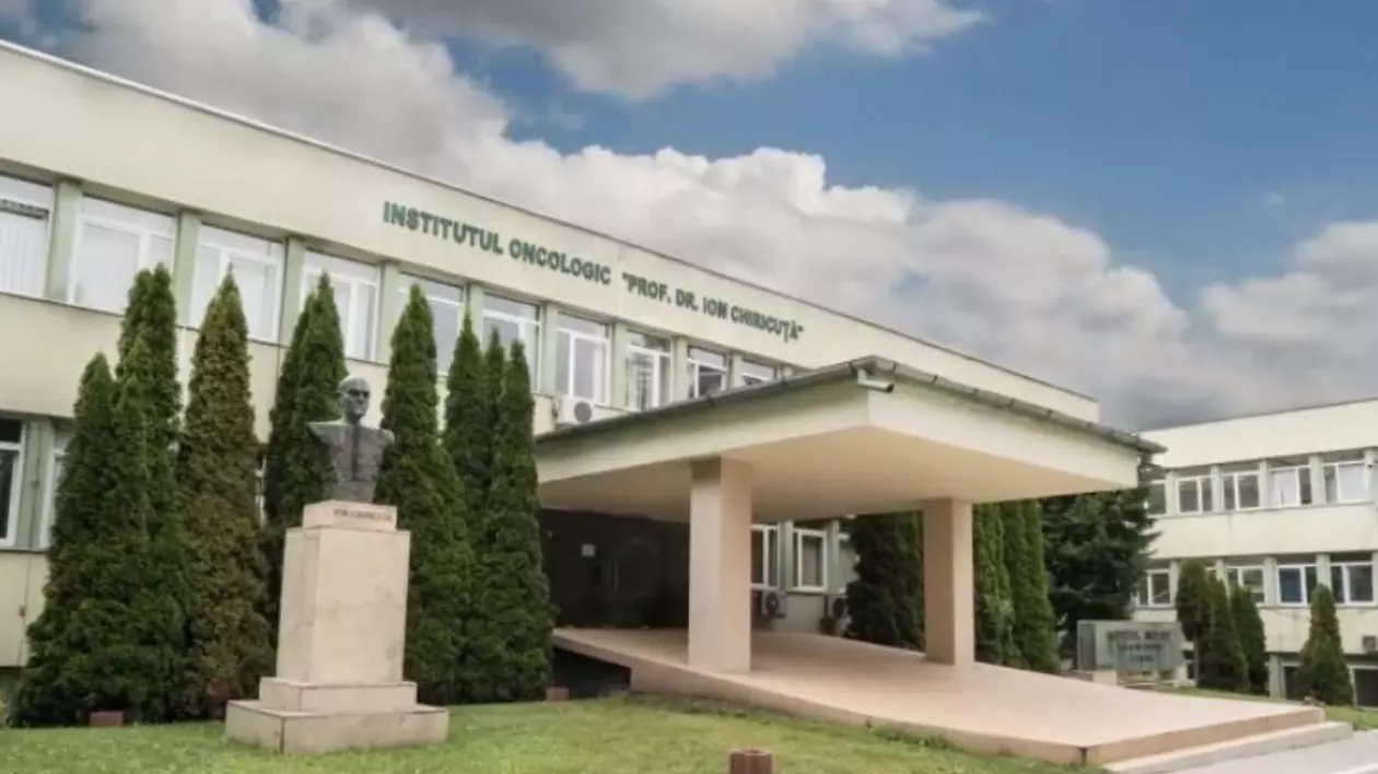 Imaginea prezintă intrarea principală a Institutului Oncologic „Prof. Dr. Ion Chiricuță” din Cluj-Napoca. Denumirea instituției este afișată deasupra intrării, iar în fundal se observă cerul acoperit de nori.