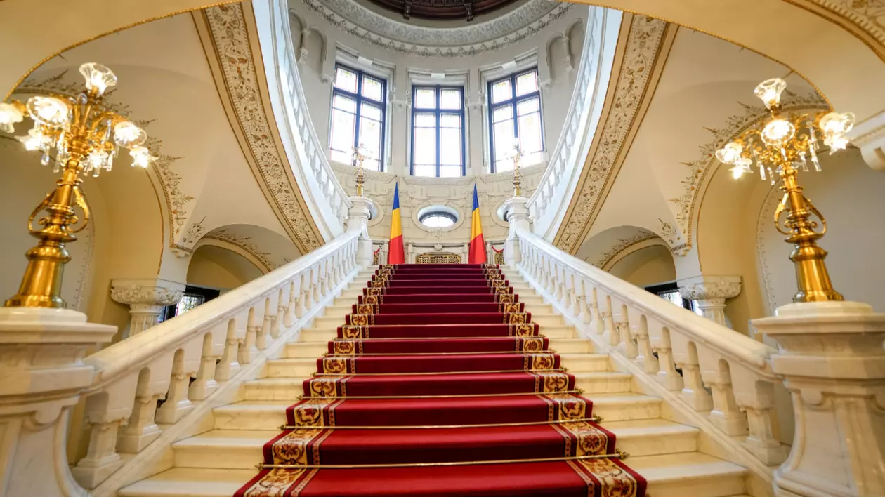 Imaginea prezintă o scară din marmură albă, acoperită cu un covor roșu, care urcă simetric spre un etaj superior luminat de un ansamblu de ferestre mari