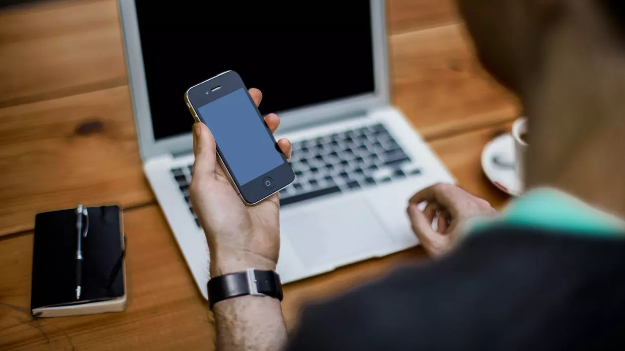 Altex: poza cu un baiat care are in fata lui pe masa un laptop, un telefon in stanga laptopului, iar in mana stanga tine un alt telegon. Masa e de lemn, iar el poarta un pluover verde