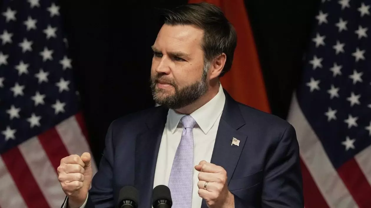 JD Vance in timp ce vorbește, mâinile ridicate și pumnii strânși, în costum închis la culoare și cravata gri deschis, cu steagul SUA în spate