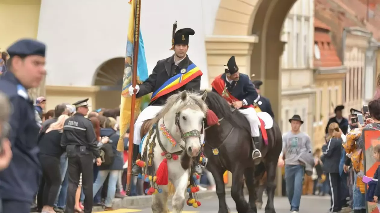 Doi bărbați îmbrăcați în costume tradiționale, defilând călare pe străzile istorice ale Brașovului. De o parte și de alta a străzii oamenii îi privesc.