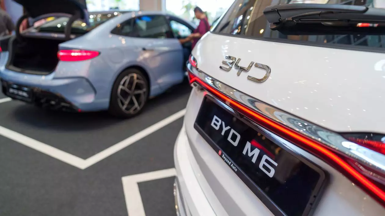 Două mașini electrice expuse într-un showroom sau la un eveniment auto. În prim-plan partea din spate a unei mașini albe marca BYD, modelul M6, evidențiat de logo-ul mare „BYD” și stopurile LED. În fundal este oa ltă mașină de culoare albă.