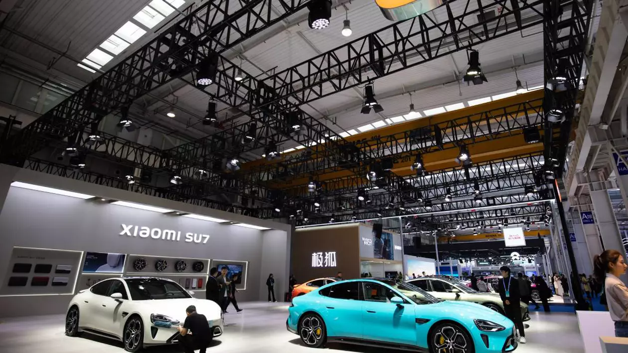 Showroom auto unde sunt expuse mai multe mașini electrice Xiaomi SU7. În prim plan sunt două modele electrice, unul alb și unul turcoaz. Printre mașini sunt cateva persoane care analizează părți ale fiecărei mașini
