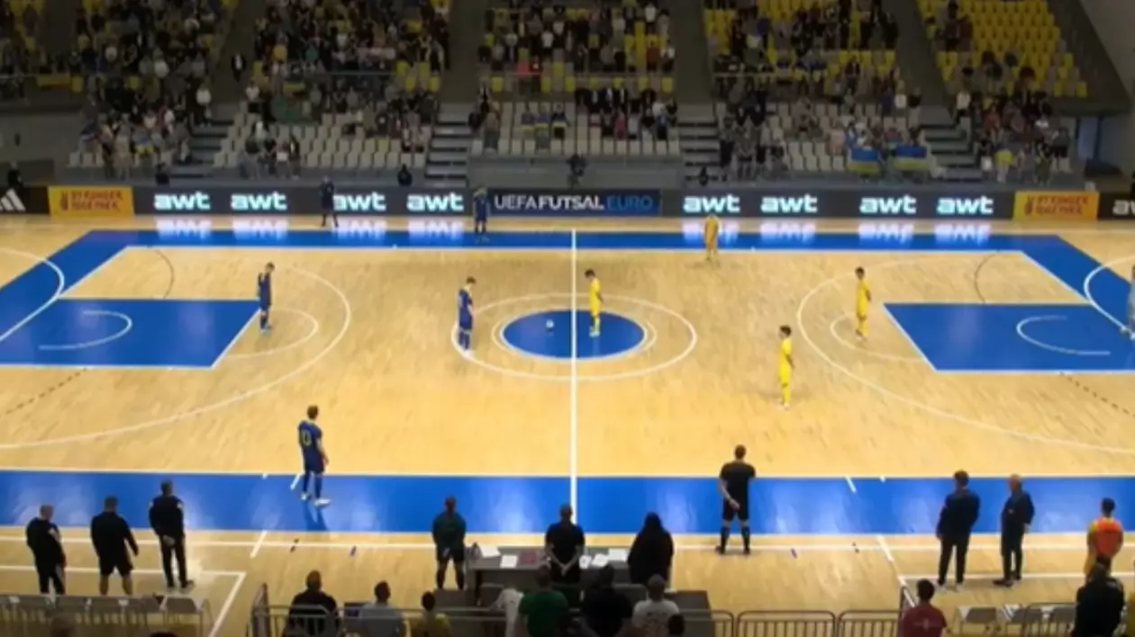 Imaginea surprinde un teren de futsal. În partea stângă sunt jucători îmbrăcați în albastru, iar în partea dreaptă sunt jucători în echipament galben. În fundal se observă tribuna cu câțiva oameni așezați pe scaune.