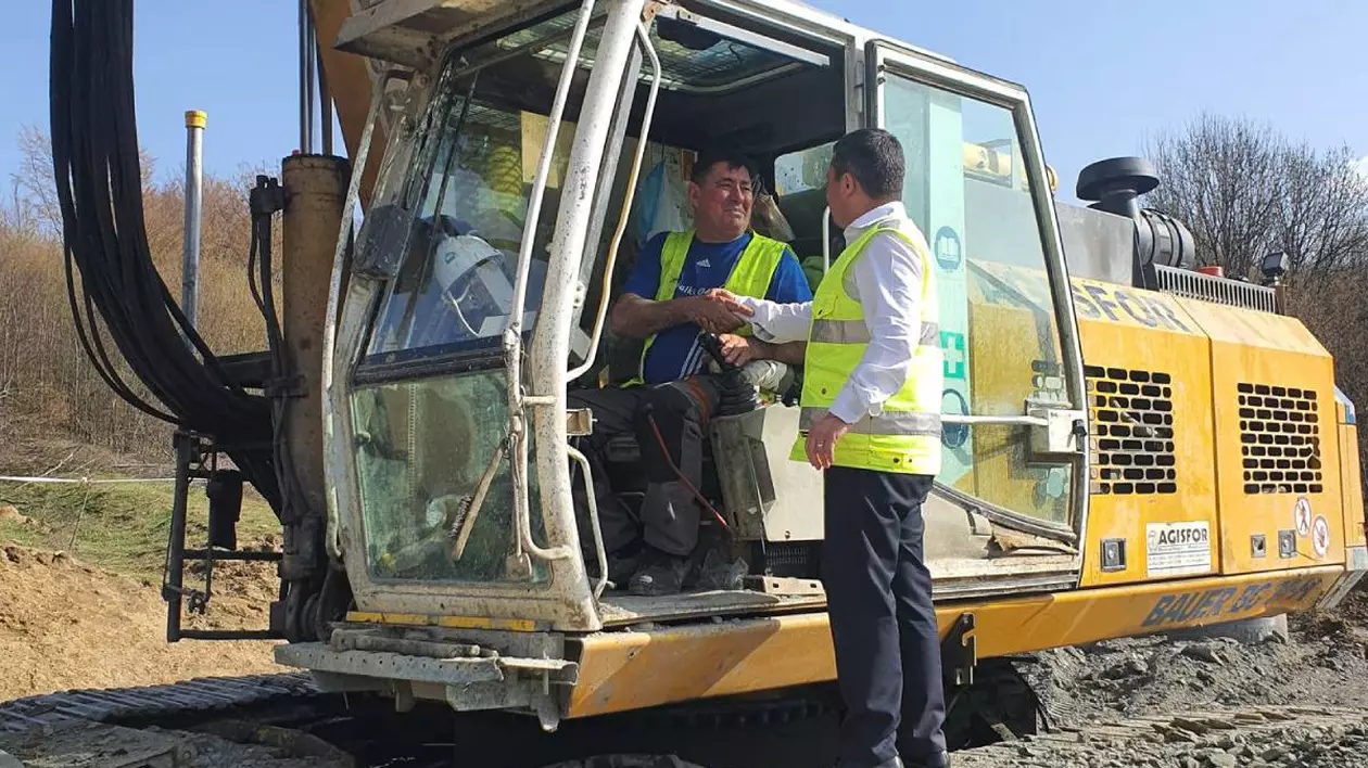 șantier de construcție pe autostrada Sibiu-Pitești, unde un muncitor aflat într-un excavator dă mâna cu un bărbat îmbrăcat în vestă reflectorizantă și cămașă albă