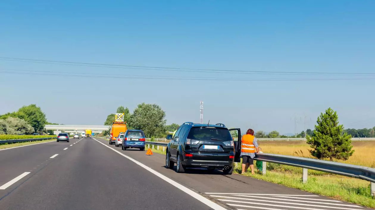 Mai multe mașini oprite pe banda de urgență de pe autostradă. Prima mașină este o mașină neagră, iar în dreapta ei este o femeie cu vestă reflectorizantă. În fața mașinii negre sunt alte două mașini oprite, care au triunghiul reflectorizant montat.