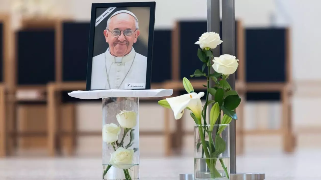 Papa Francisc a murit, imagine cu o fotografie cu Papa pe un pahar cu flori alve, alaturi un alt pagar cu trandafiri albi iar in spate niste scaune negre