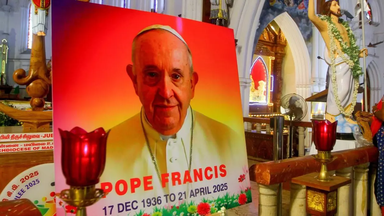 Un portret al Papei Francisc, cu scris pe el și o candelă aprinsă, o statuie a lui Isus Cristos în spate, într-o biserică