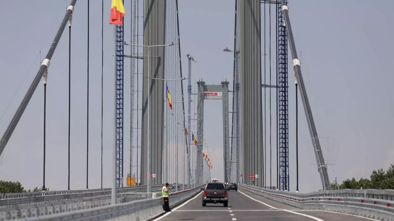 podul suspendat peste Dunăre de la Brăila, cu mașini care circula pe drum și stegulețe tricolore arborate de-a lungul cablurilor de susținere, fără trafic intens
