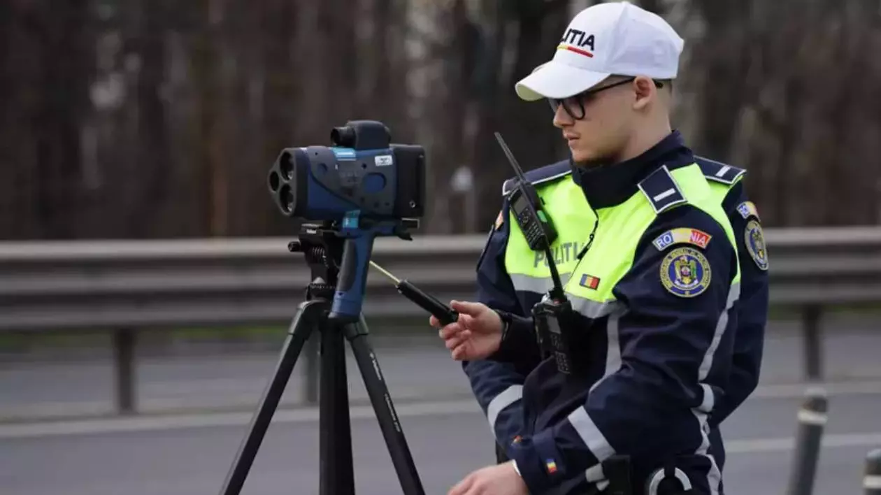 Polițist rutier echipat cu uniforma specifică și o șapcă albă inscripționată cu „POLIȚIA”. Acesta operează un radar mobil de tip pistol montat pe un trepied. Pe fundal se observă un drum național, delimitat de un parapet metalic.