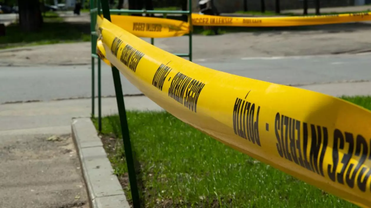 În prim-plan se vede o bandă galbenă, cu inscripția „Poliție, acces interzis”, cu care este împrejmuită o zonă verde de pe o stradă.