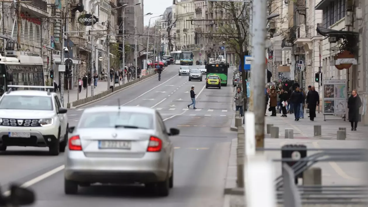 O zi cu nori, în care mai multe mașini și troleibuze circulă pe stradă, câțiva oameni sunt pe trotuar, iar un bărbat traversează strada pe trecerea de pietoni, București, Piața Universității, aprilie 2025