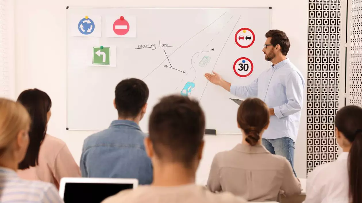 elevi intr-o sala de clasa, in fata lor se afla un profesor care arata spre tabla, unde sunt cateva indicatoare rutiere