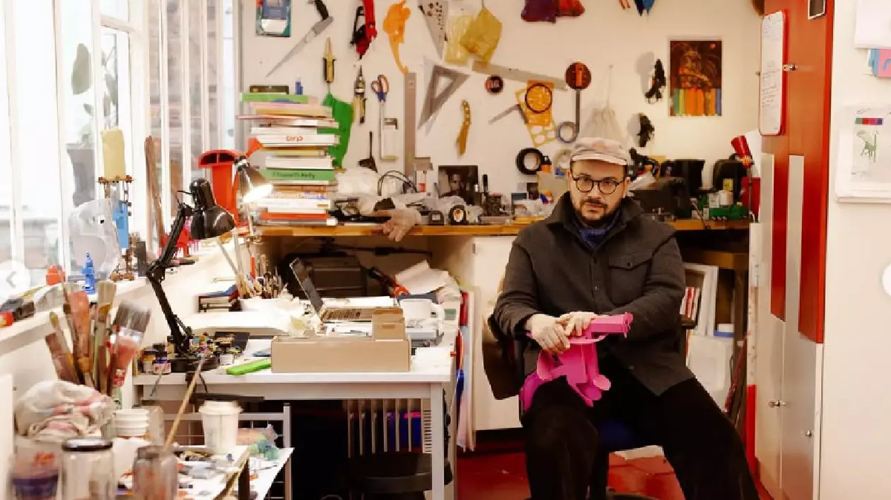 Un bărbat într-o studio aglomerat, multe unelte pe pereți, un birou pe care se află un laptop, o lampă și multe alte obiecte