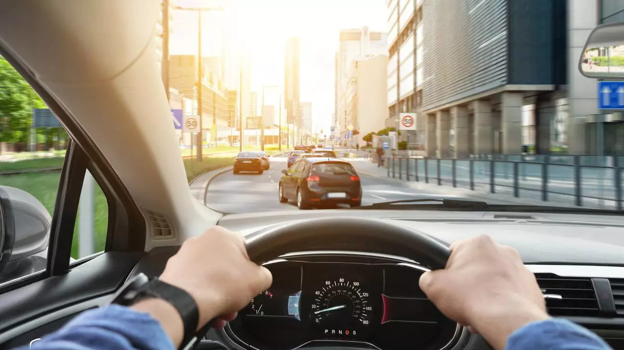 Șofer aflat la volanul unei mașini aflate în trafic. Șoferul ține ambele mâini pe volan. În față se observă mai multe vehicule pe o șosea cu două benzi, clădiri moderne de birouri și indicatoare rutiere