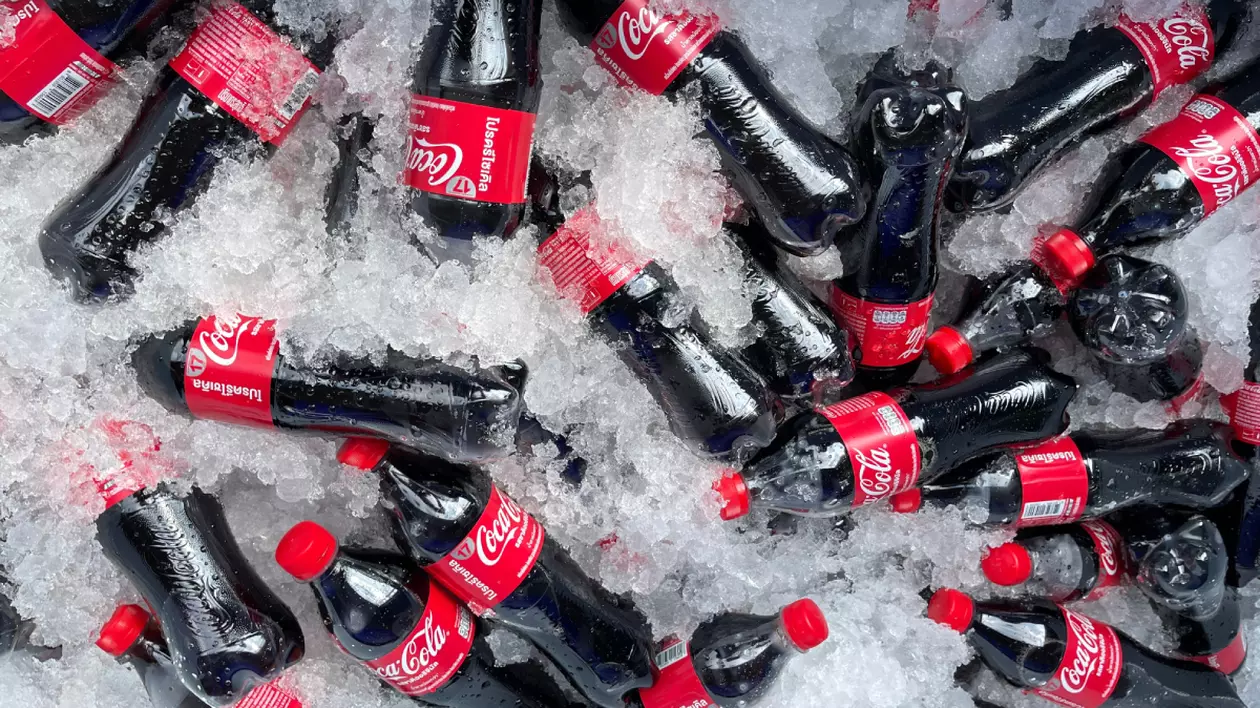 Mai multe sticle de suc Coca-Cola așezate printre cuburi de gheață