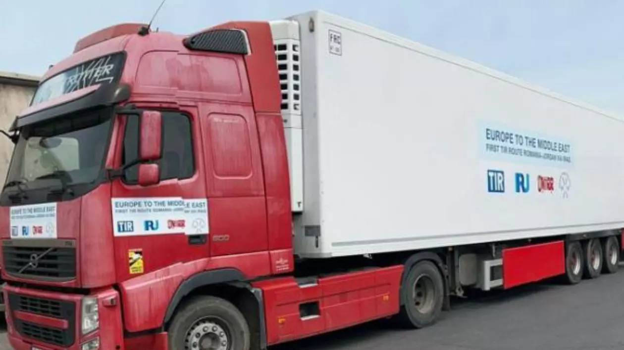 camion de transport marfă, tir, cu cap tractor roșu și remorcă albă, parcat pe marginea unui drum. Pe lateralul remorcii este lipit un afiș cu textul „ europe to the middle east”