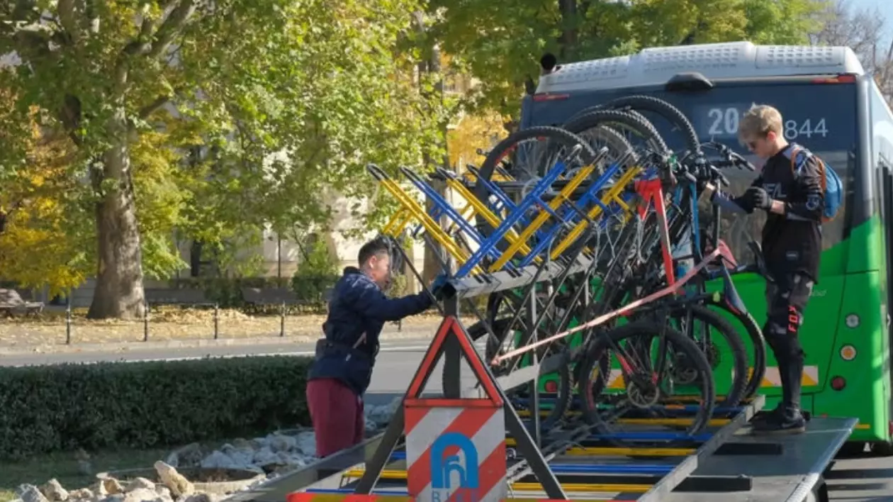 În imagine se observă un sistem de transport al bicicletelor montat în spatele unui autobuz, într-un peisaj urban. Două persoane, îmbrăcate în echipament sportiv, sunt ocupate cu încărcarea sau descărcarea bicicletelor de pe suportul special.