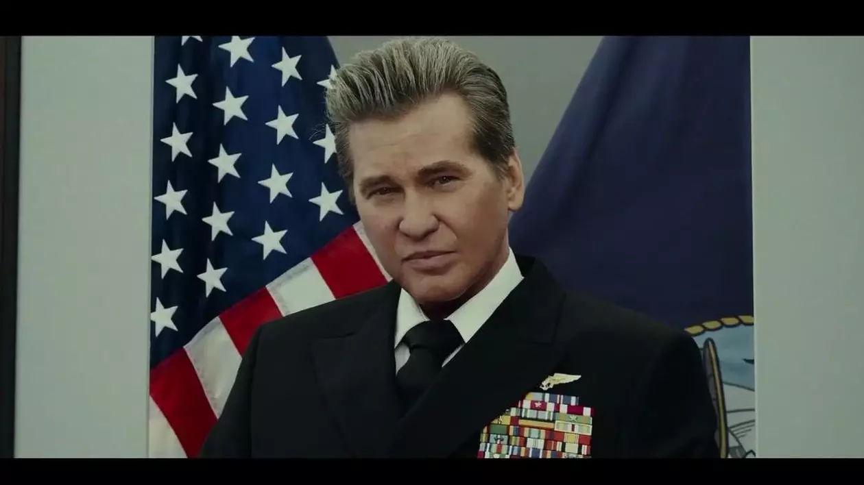 Val Kilmer în rolul lui Ice, în filmul Top Gun: Maverick, îmbrăcat în uniformă, cu steaguri în spate