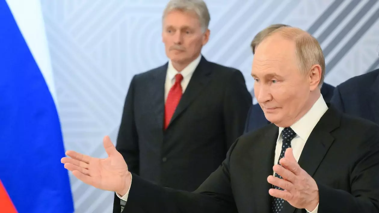 Putin cu brațele deschise, îmbrăcat în costum negru, camasa alba si cravata neagra. În spate, Dmitri Peskov, secretarul de presă al Kremlinului, în costum negru, cîmașă albă și cravată roșie. Președintele rus Vladimir Putin în timpul întâlnirii sale cu președintele egiptean Abdel Fattah El-Sisi în marja celui de-al 16-lea Summit BRICS de la Kazan.