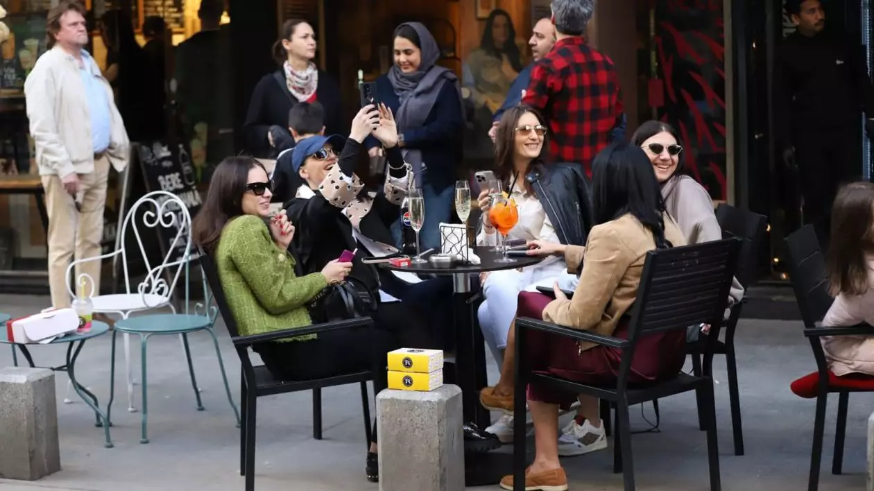 Un grup de cinci tinere stau la o masă, vorbesc, râd și fac poze, iar în jurul lor sunt alți oameni, în Centrul vechi, București, 13 aprilie 2025