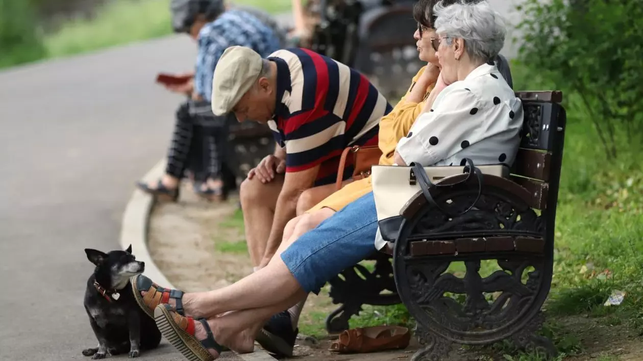 Două femei în vârstă stau de vorbă pe o bancă, iar lângă ele este un bărbat. În fața lor este un cățel care stă jos, iar în spate se vede iarba verde. Lângă banca lor, este o altă bancă pe care sunt oameni, parcul Cișmigiu, București, 25 aprilie 2025
