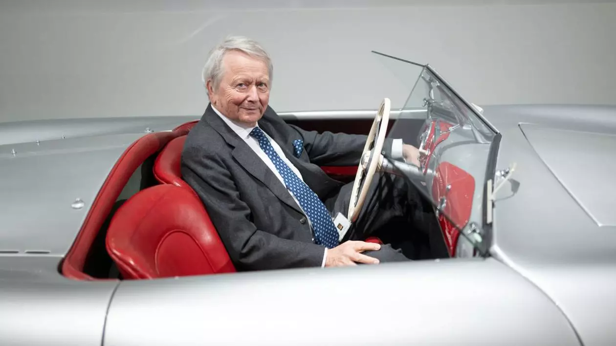 Wolfgang Porsche îmbrăcat într-un costum gri și cravată albastră, așezat la volanul unui automobil decapotabil de epocă, cu interior roșu