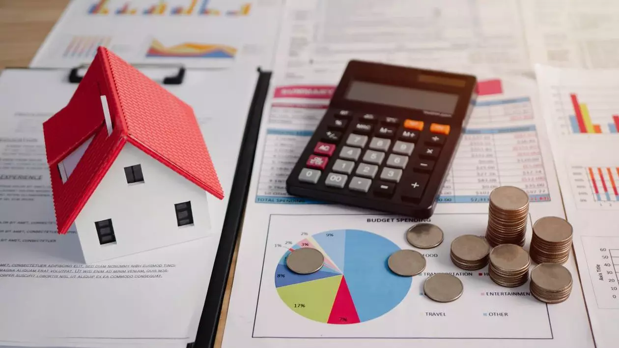 În stânga imaginii este o hârtie scrisă pe care stă o căsuță în miniatură cu acoperiș roșu, iar în dreapta se vede o pagină cu grafice colorate, un calculator negru pe ea și niște monede