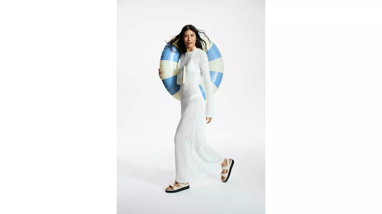 Colecție MODIVO Summer 2025. Model, fata cu parul lung si brunet, imbracata intr-un caftan de plasa alb, lung, cu manecu lungi, tine pe umar un colac de piscina mare, alb, cu dulgi albastru deschis