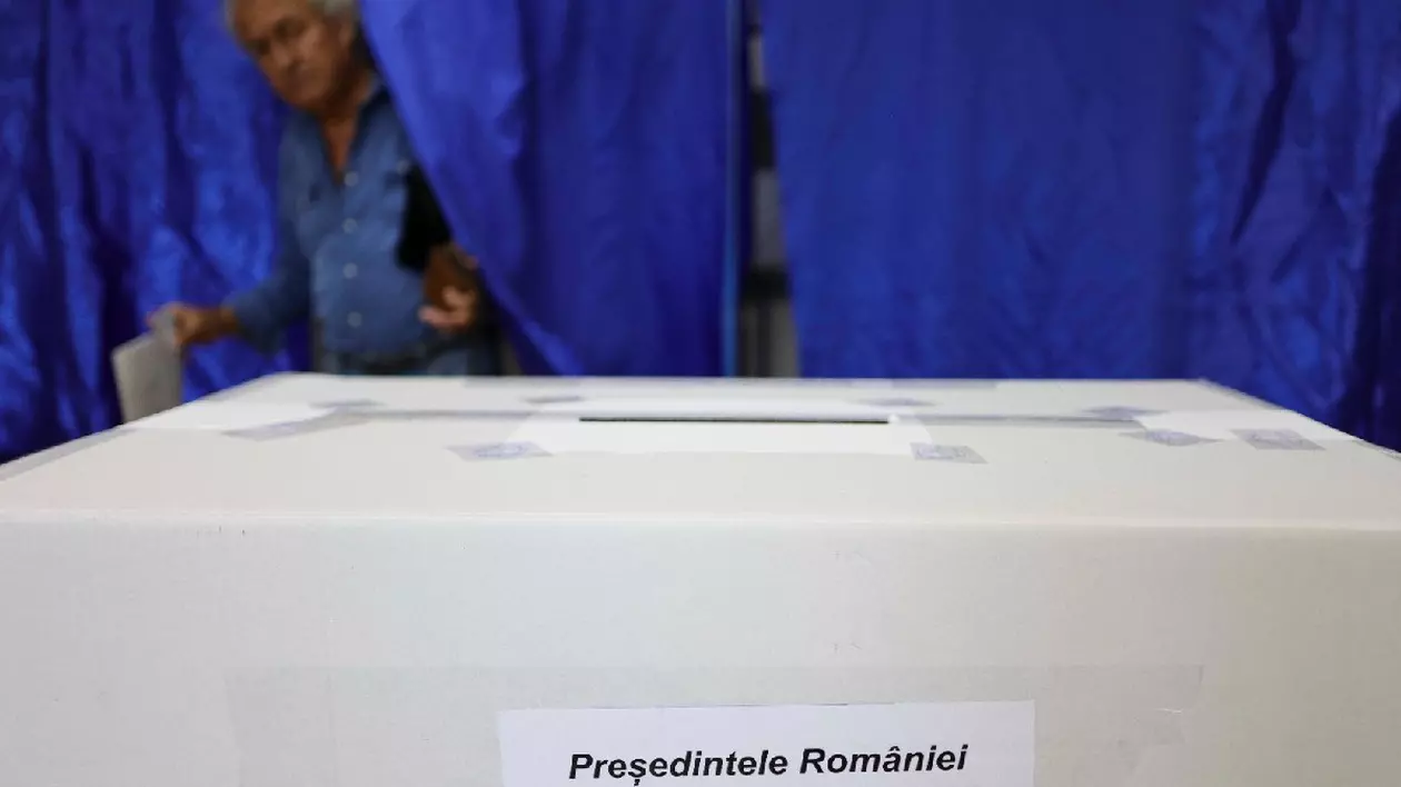 Un bărbat îmbrăcat în cămașă albastră, iese dintr-o cabină de vot, în față se vede o urnă albă, pe care scrie „Președintele României”
