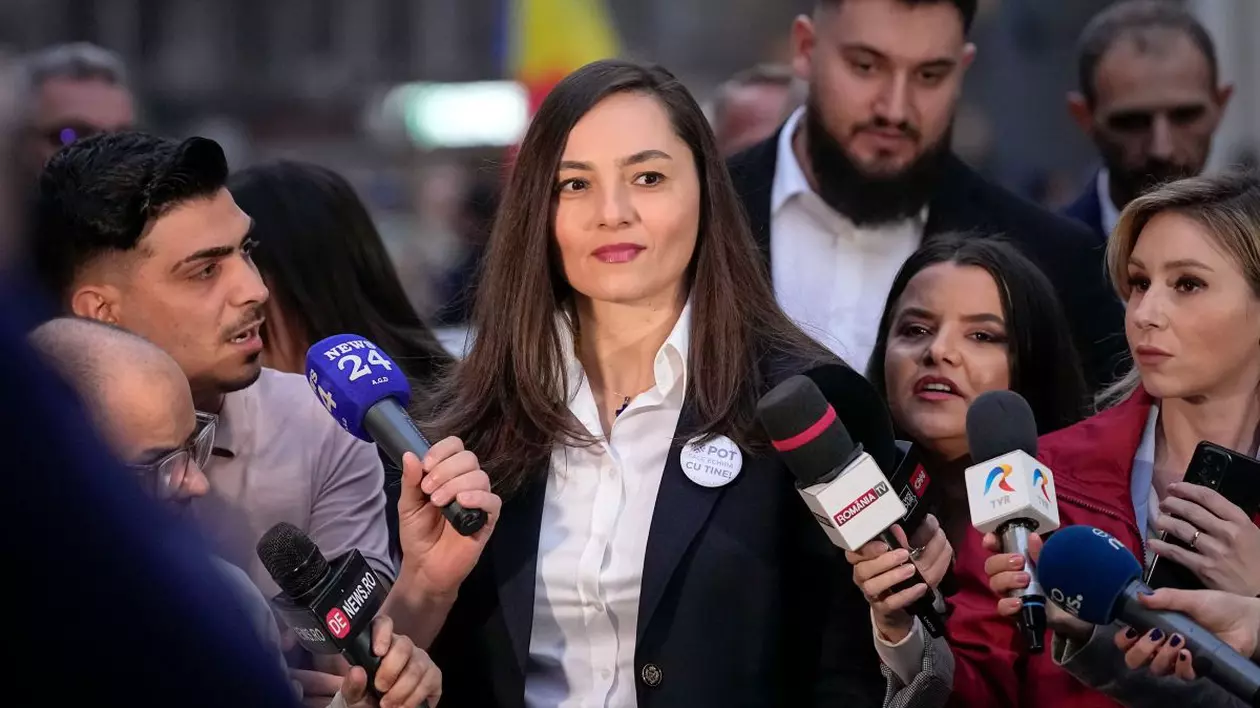 Anamaria Gavrilă, îmbrăcată cu un sacou și o cămașă albă, în timp ce este înconjurată de jurnaliști cu microfoane, care îi pun întrebări, în fața Biroului Electoral Central