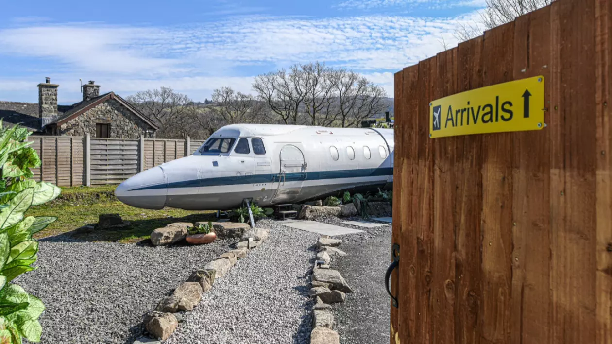 În imagine este ilustrat un avion vechi, aflat într-o grădină. Pe jos este mult pietriș, iar cerul este senin