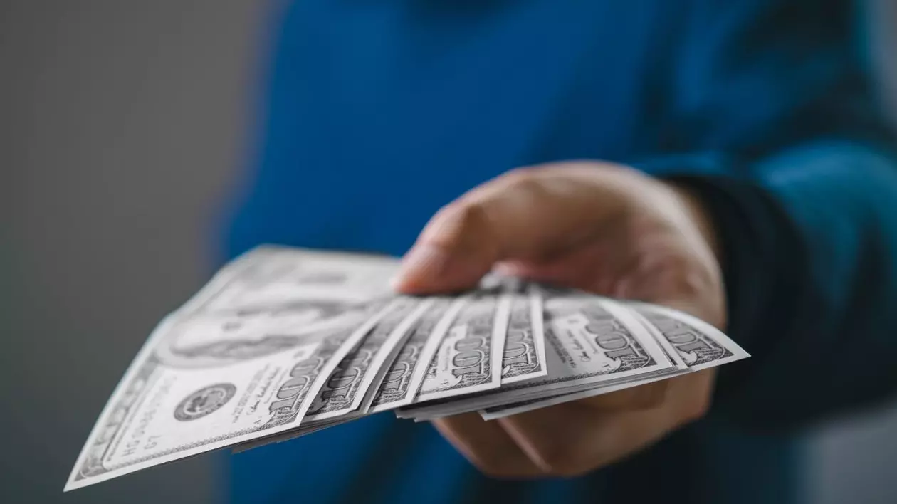 O imagine de aproape a unei mâini care oferă mai multe bancnote de dolari americani, semnificând o tranzacție financiară, un împrumut sau o plată într-un context de afaceri