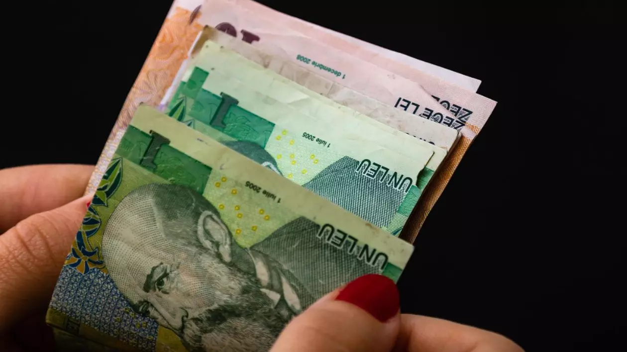 Prim plan cu mâna unei femei, cu ojă roșie pe unghii, care numără mai multe bancnote de un leu și 10 lei, pe fundal negru
