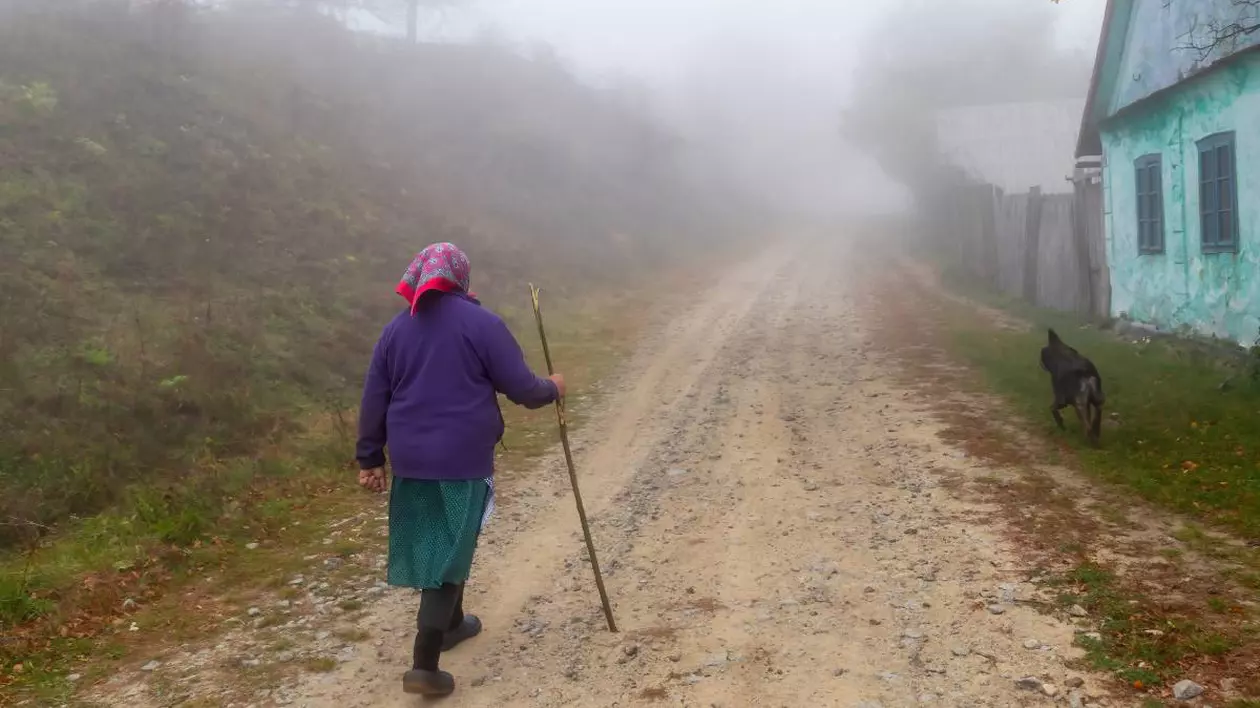 O bătrână de aproximativ 82 de ani, îmbrăcată gros, se plimbă în baston pe un drum de țară, pietruit.