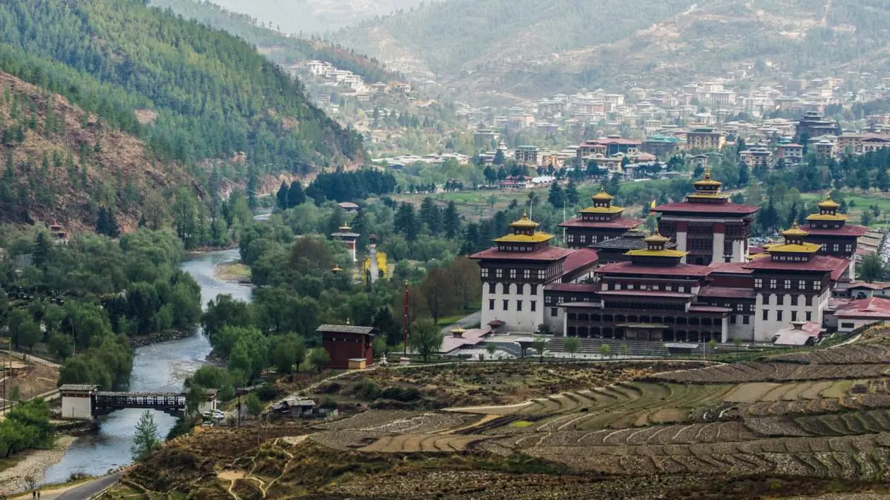 Câmpuri de orez pregătite pentru plantare în fața în Thimphu, Bhutan, imagine cu o asezare între munti impaduriti , cu temple si case in departare si o apă care curge la vale, in spate putina ceață