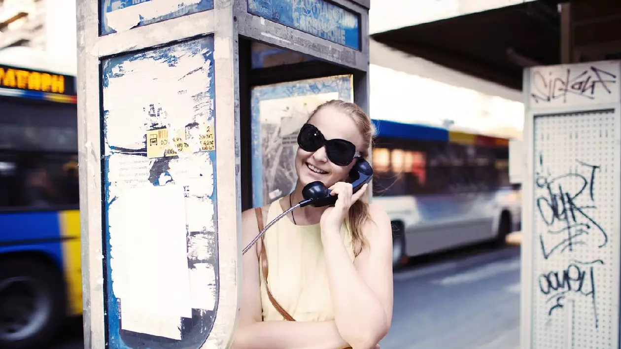 Femeie atrăgătoare cu ochelari de soare, în vacanța de vară, vorbind la un telefon public într-o cabină de-a lungul unei străzi urbane, în timp ce autobuzul trece prin spate