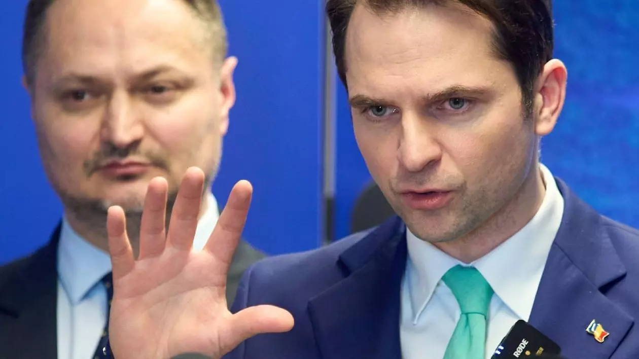 Cadru foarte strâns cu Sebastian Burduja, ministrul economiei, care ține o conferință îmbrăcat în sacou negru, cu o cravată bleu deschis și cămașă albă, cu mâna dreaptă ridicată și palma deschisă