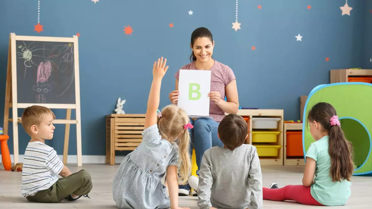 Un grup de copii înscriși deja la creșă sau la grădiniță, în tip ce învață alfabetul de pe o planșă pe care le-o arată educatoarea.
